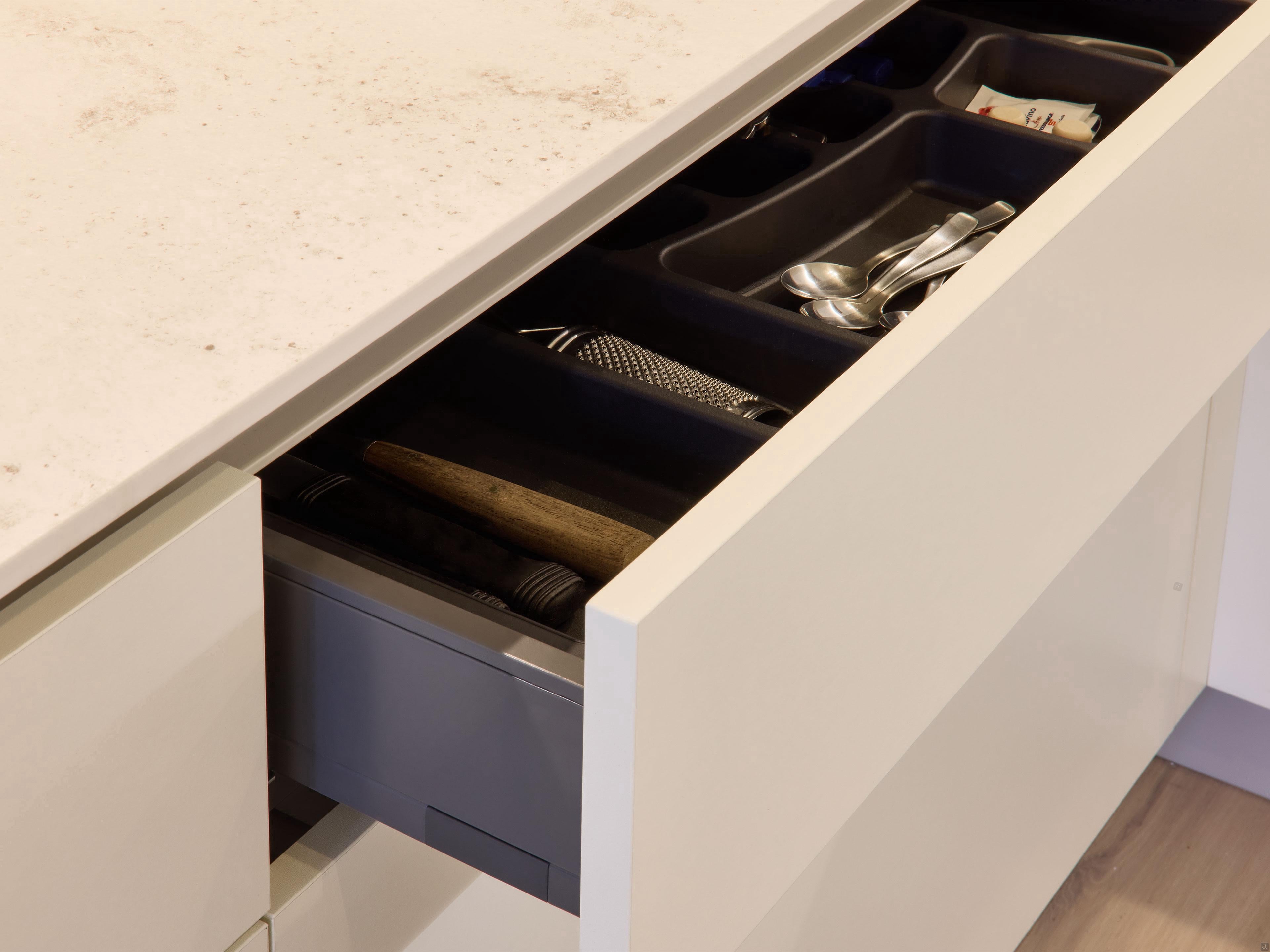A partially open drawer from Modern Kitchens by Kitchen Furniture reveals organized cutlery in compartments under a light countertop, all within sleek white cabinetry that highlights a modern kitchen design.