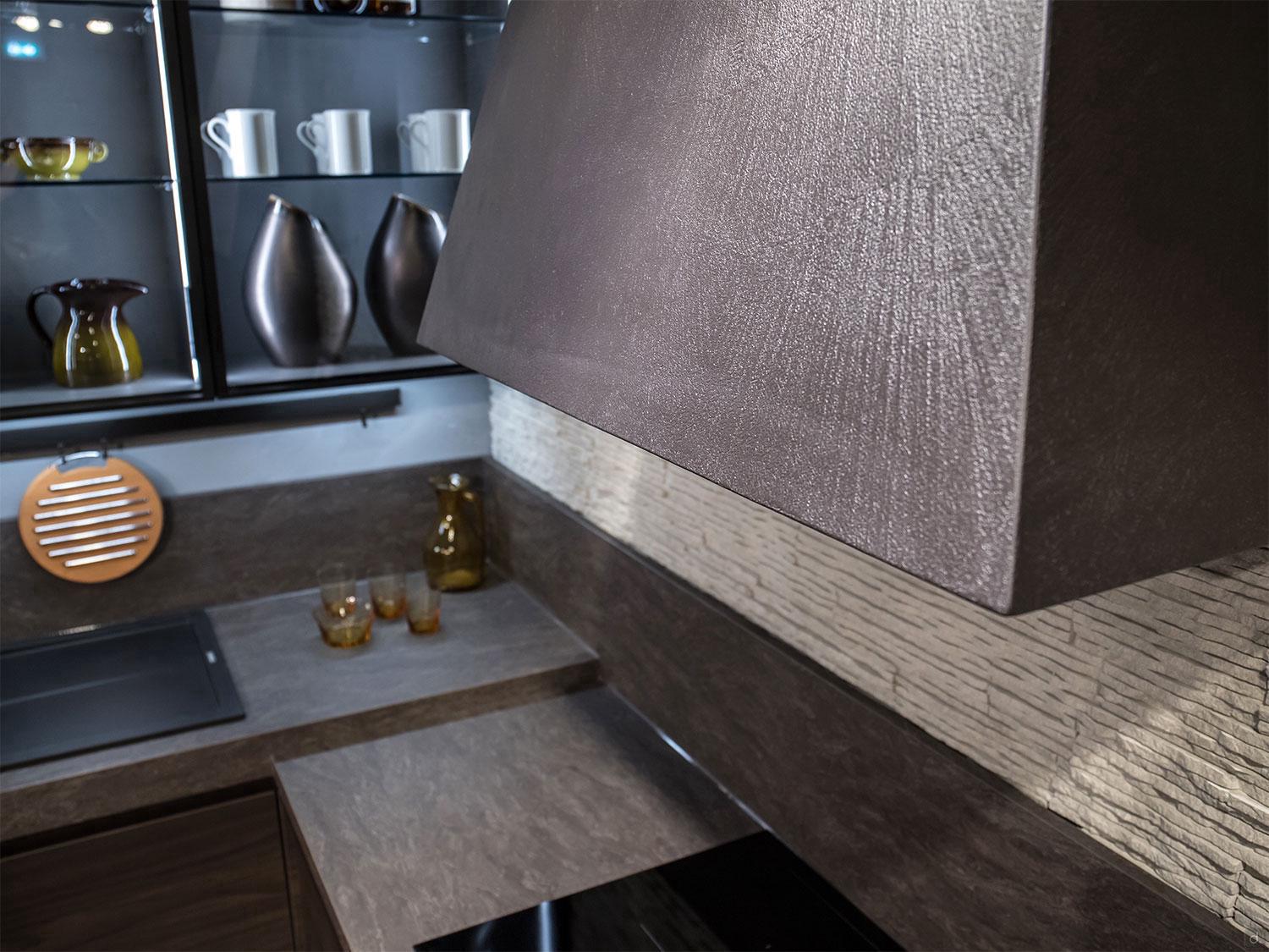 The Urban Kitchens by ORDER Kitchen Furniture features a metallic range hood, brown countertops, custom cabinetry, glassware and ceramics on open shelves, and a round wooden utensil holder on a textured backsplash.