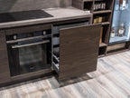 The Urban Kitchens by ORDER Kitchen Furniture features a modern kitchen with custom cabinets, built-in oven, electric cooktop, partially open wooden drawer, wood flooring, and shelves displaying glassware and bottles.