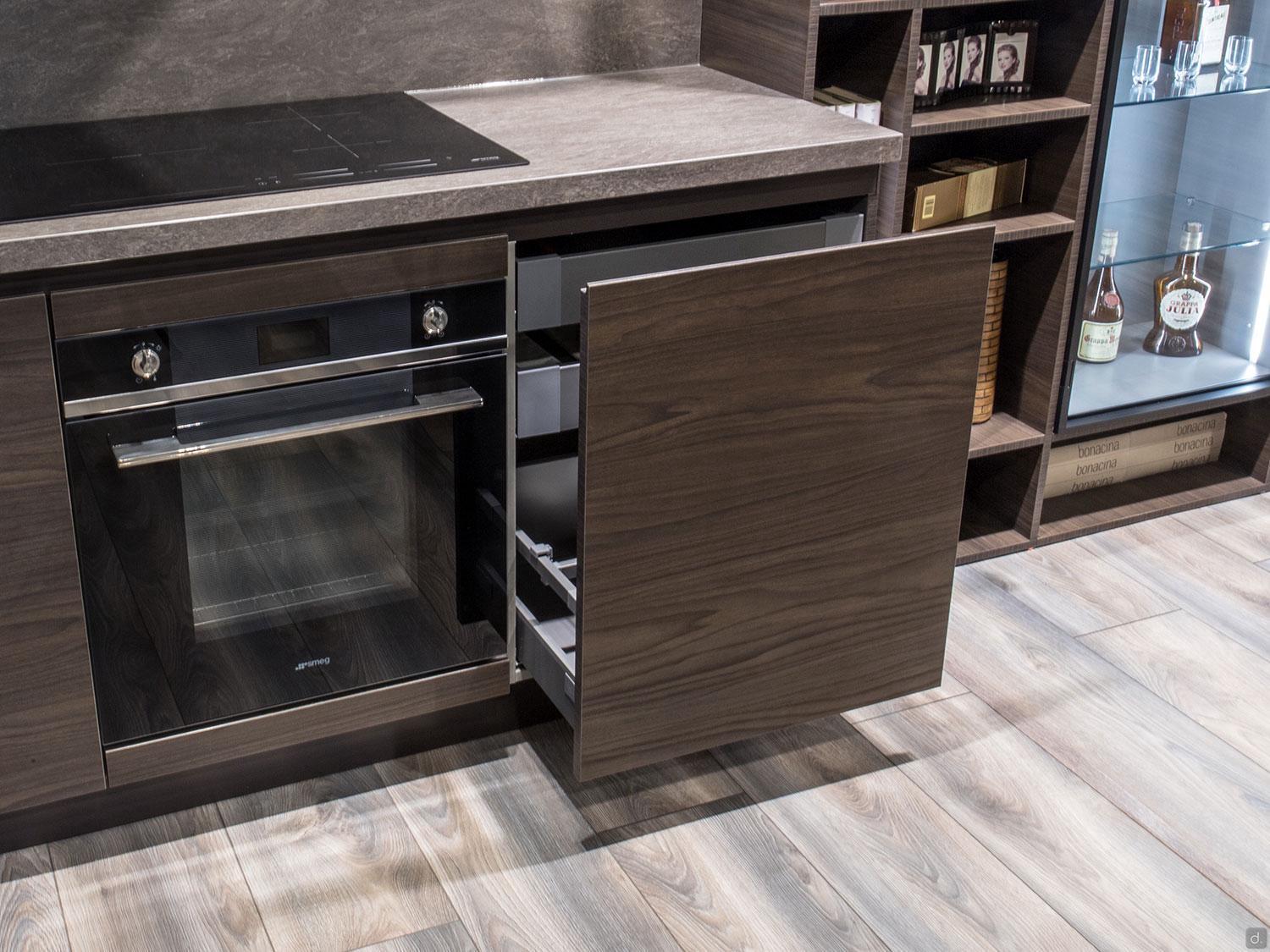 The Urban Kitchens by ORDER Kitchen Furniture features a modern kitchen with custom cabinets, built-in oven, electric cooktop, partially open wooden drawer, wood flooring, and shelves displaying glassware and bottles.