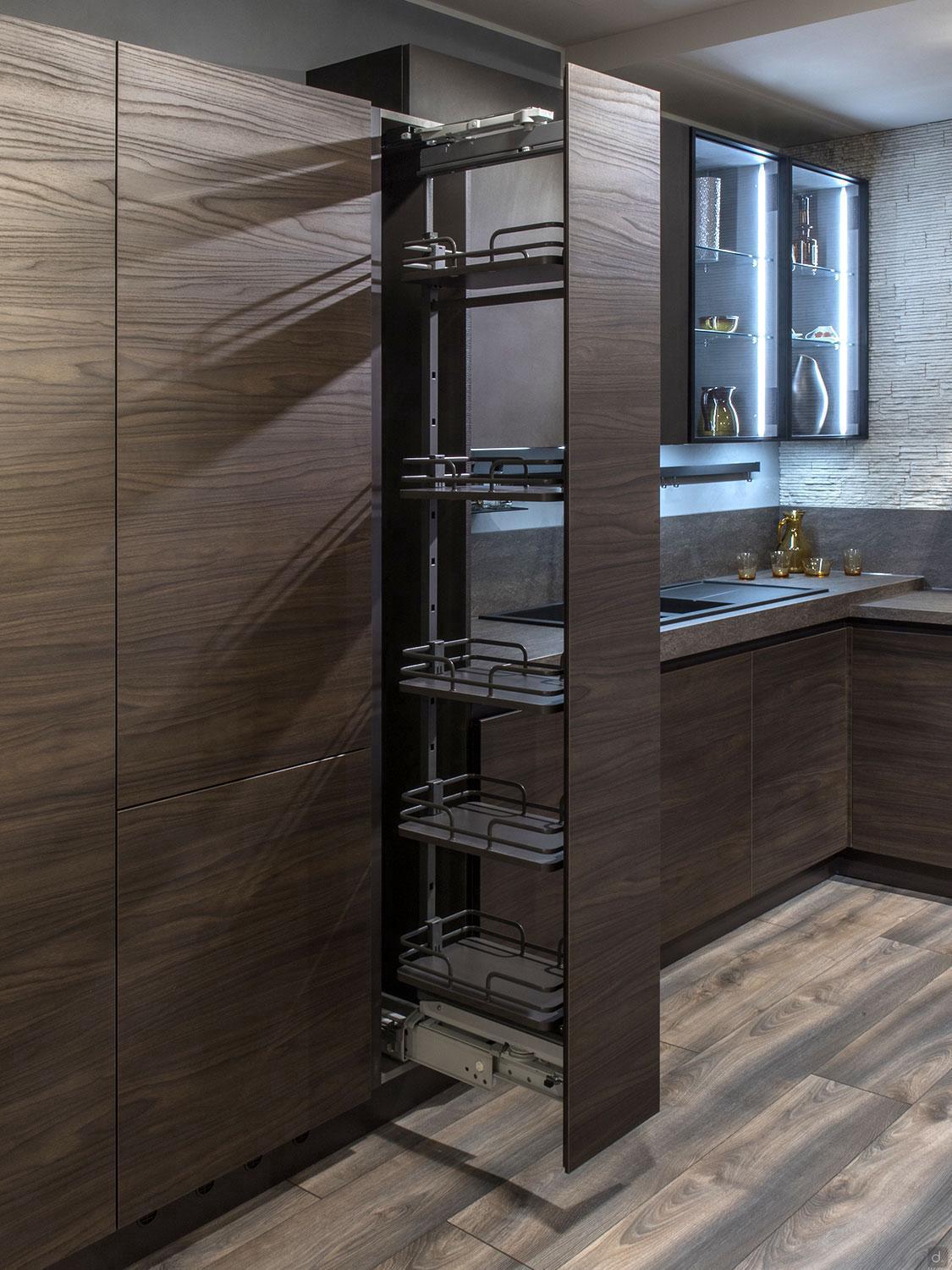 A modern kitchen by ORDER Kitchen Furniture’s Urban Kitchens features dark cabinets and a tall, slim pull-out pantry with metal shelves, next to countertops and glass-front uppers displaying décor—custom furniture at its finest.