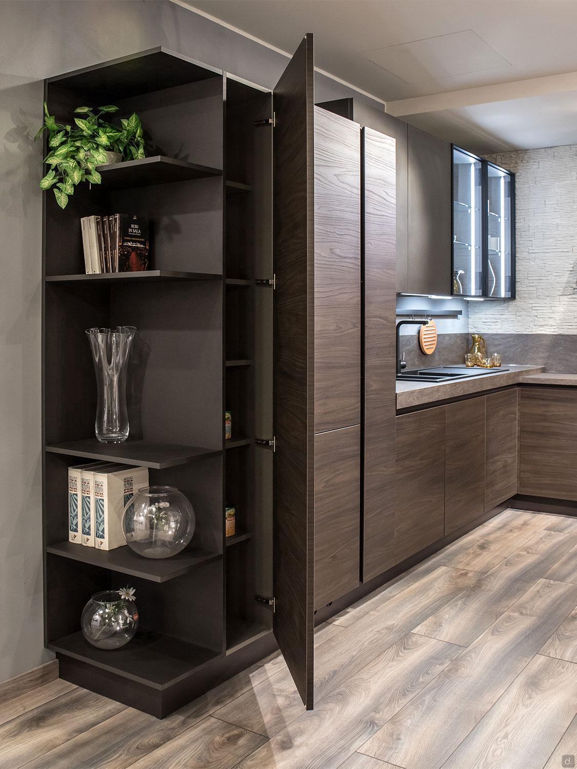 Urban Kitchens by ORDER Kitchen Furniture combines dark wood cabinets, custom pieces, and open shelving for decor and glassware. A tall cabinet conceals extra storage, while light wood floors and greenery add a warm, modern touch.