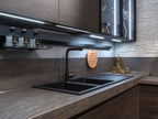 Urban Kitchens by ORDER Kitchen Furniture features a modern space with dark cabinets, custom furniture, a double black sink, under-cabinet lighting, and stone backsplash. Glassware is neatly arranged and a wooden trivet hangs above the sink.