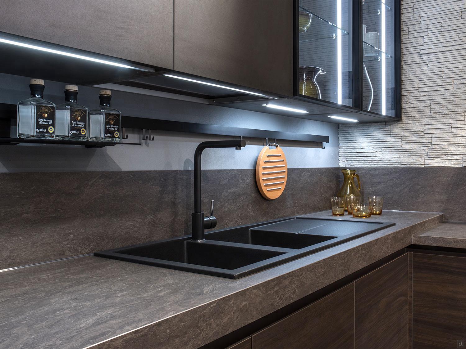 Urban Kitchens by ORDER Kitchen Furniture features a modern space with dark cabinets, custom furniture, a double black sink, under-cabinet lighting, and stone backsplash. Glassware is neatly arranged and a wooden trivet hangs above the sink.