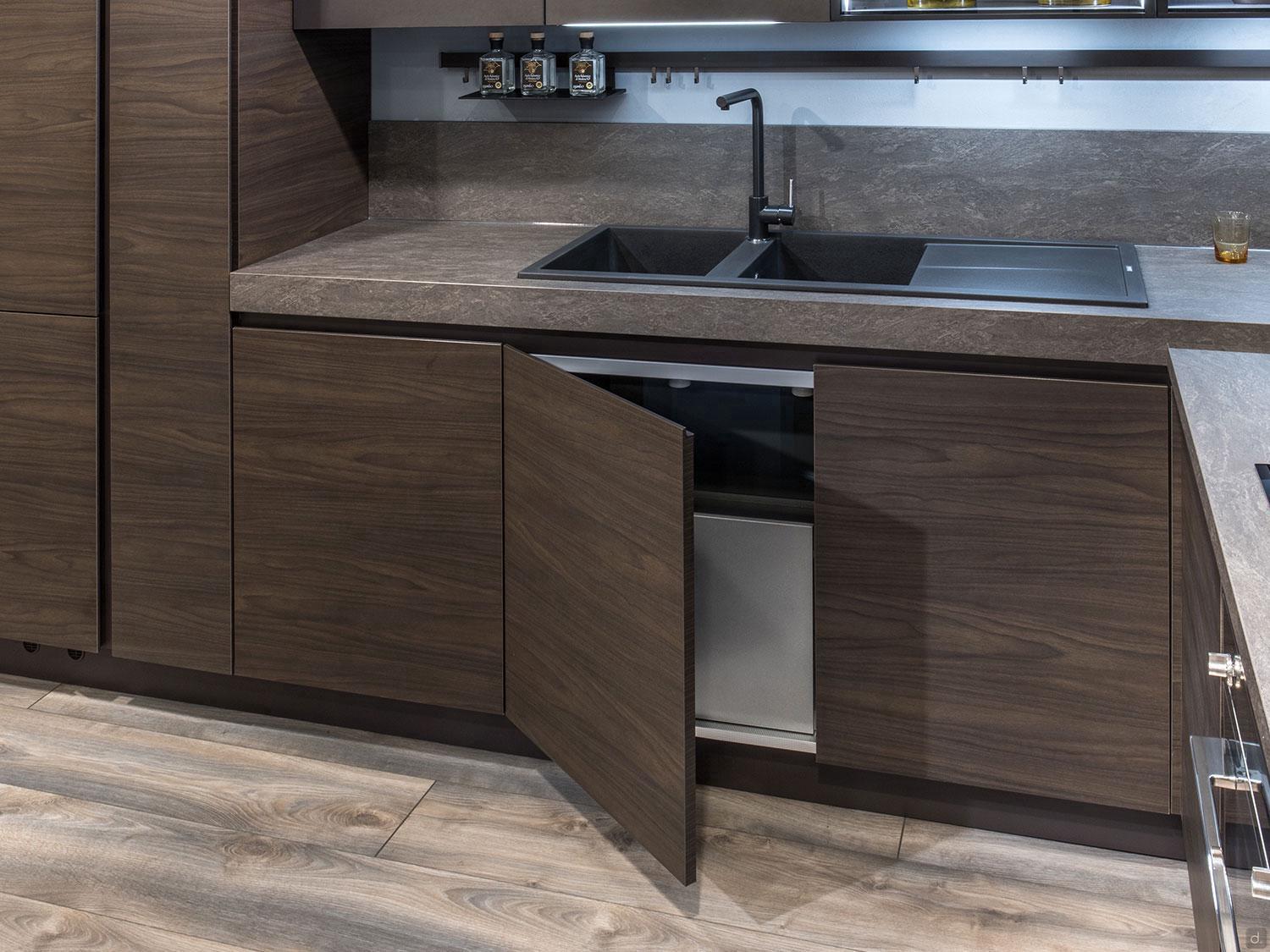 An Urban Kitchens setup by ORDER Kitchen Furniture features dark wood cabinetry, a partially open lower door, black sink and faucet, gray countertops, bottles on a shelf above the sink, and a drink placed on the counter.