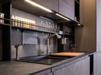 Discover Urban Kitchens by ORDER Kitchen Furniture: a modern Yerevan-style space with dark cabinets, industrial features, under-cabinet lighting, built-in sink, sleek faucet, spice jars, glassware shelves, and a cutting board on a black countertop.