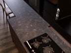 The Urban Kitchens set by ORDER Kitchen Furniture features a dark marble countertop, built-in black gas stovetop with three burners, wooden chairs, and a wood-like floor for a sleek, modern industrial look.