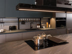 The Urban Kitchens by ORDER Kitchen Furniture features dark cabinets, custom units, a built-in oven, black sink, and sleek stovetop. Toaster, spice rack, and utensils are organized on the counter with warm lighting for a cozy feel.