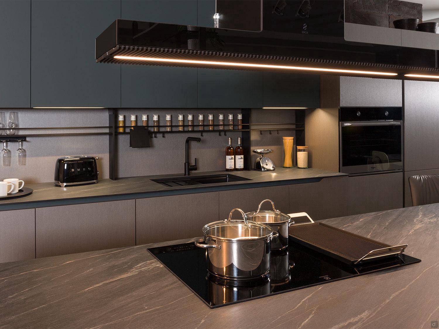 The Urban Kitchens by ORDER Kitchen Furniture features dark cabinets, custom units, a built-in oven, black sink, and sleek stovetop. Toaster, spice rack, and utensils are organized on the counter with warm lighting for a cozy feel.