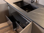 Urban Kitchens by Kitchen Furniture features a modern sink with a black faucet and integrated drainboard in a gray countertop. Two open drawers below offer organized storage and separate compartments for trash and recycling bins—ideal for urban kitchens.