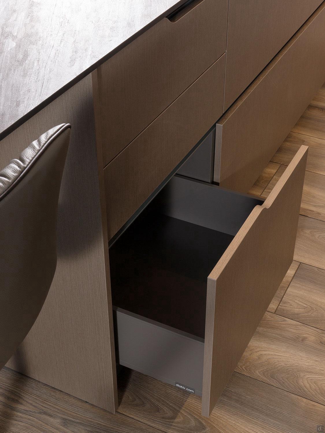 A close-up of Kitchen Furniture's Urban Kitchens cabinet on a light wood floor, featuring an open bottom drawer with a sleek black interior, positioned next to a cushioned chair.