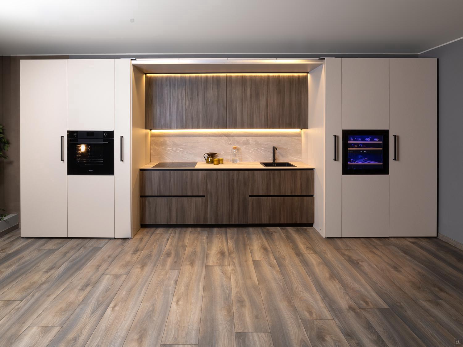 Urban Kitchens by Kitchen Furniture features light wood floors, beige cabinets, a built-in black oven, wine cooler, and a central cooking area with a wood-paneled backsplash and under-cabinet lighting—ideal for modern urban spaces.