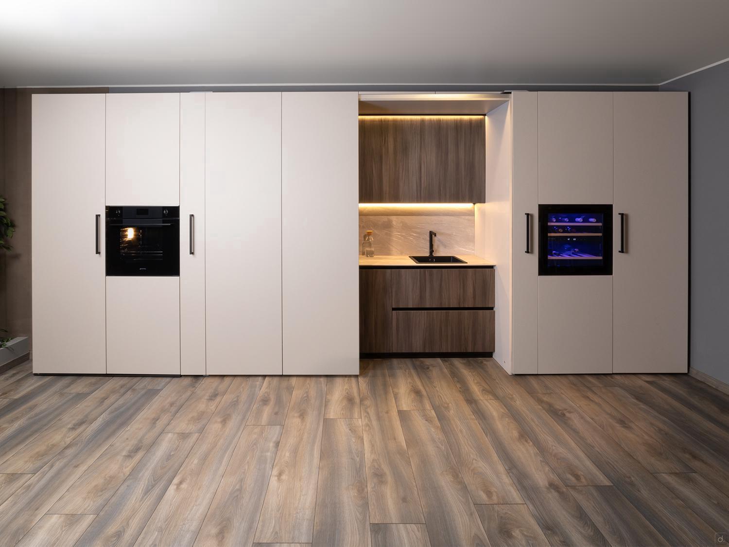 The Urban Kitchens collection by Kitchen Furniture features a modern minimalist design with light beige cabinets, built-in black oven/microwave, wood-textured lowers, small sink area, under-cabinet lighting, and light wood flooring—ideal for city homes.