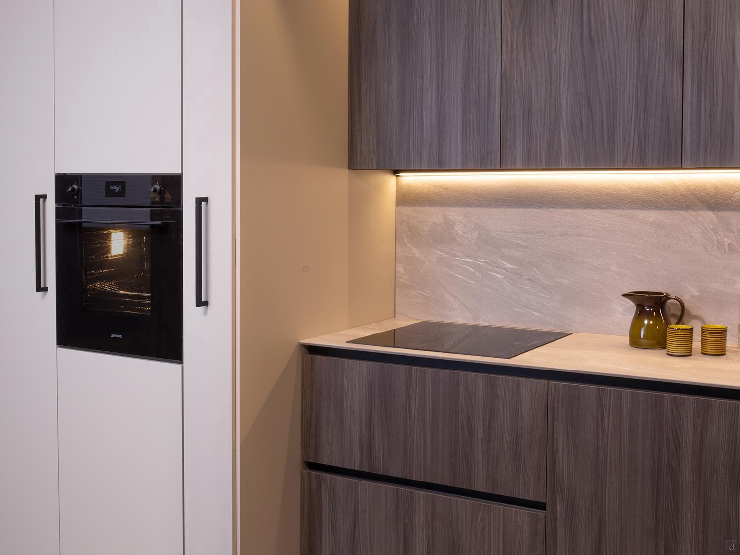 Urban Kitchens by Kitchen Furniture features dark wood cabinets, a built-in black oven on the left, beige countertop, induction cooktop, and decorative accents like a brown pitcher and yellow cups under under-cabinet lighting.